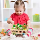 A young toddler girl sitting on a rug and playing with a wooden carrot harvest toy, practicing fine motor skills and hand-eye coordination.