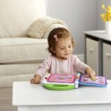 A toddler girl interacting with the LeapFrog bilingual toy book at a table, showing the touch-sensitive pages that teach food, animals, and colors in two languages.